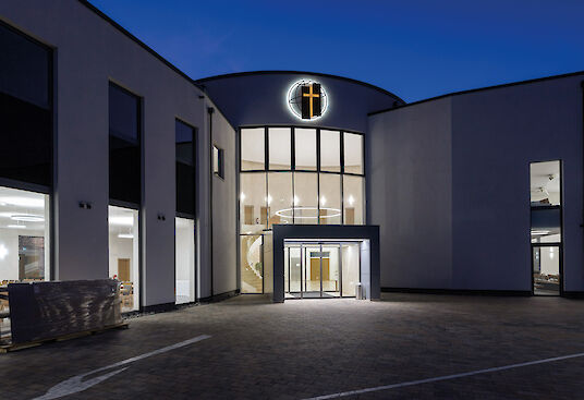 Salón parroquial de Schwegenheim de noche, entrada iluminada y cruz con acento luminoso sobre el portal.