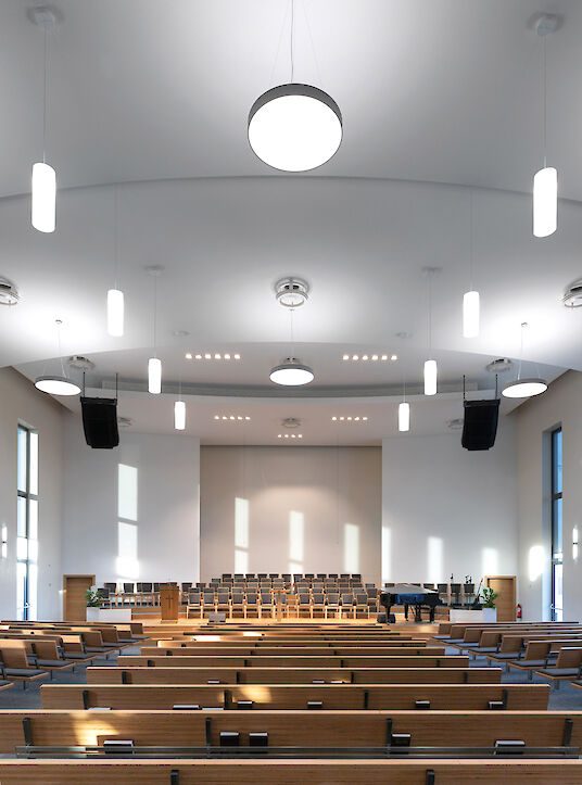 Salón de la iglesia con asientos de madera y luminarias suspendidas redondas «Basic» en un techo abovedado.
