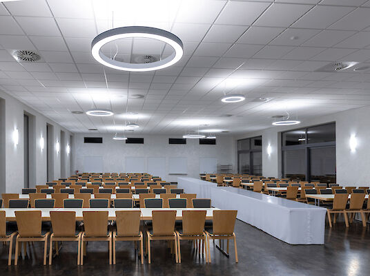 Veranstaltungssaal des Gemeindehaus Schwegenheim mit Holzstühlen und ringförmigen Ringo Star Pendelleuchten.