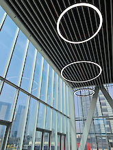 Kreisförmige, leuchtende Pendelleuchten in unterschiedlichen Größen hängen unter einer hohen Glasdecke mit schmalen, parallel verlaufenden Metallstreben und beleuchten den modernen Innenraum.