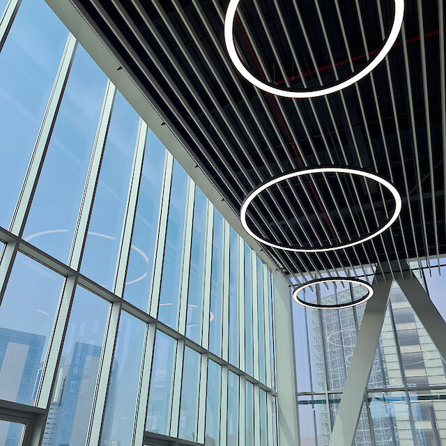 Kreisförmige, leuchtende Pendelleuchten in unterschiedlichen Größen hängen unter einer hohen Glasdecke mit schmalen, parallel verlaufenden Metallstreben und beleuchten den modernen Innenraum.