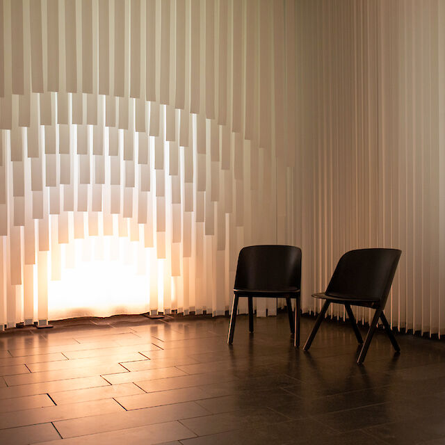 Two black chairs stand in front of a wall of vertical white louvres arranged in an undulating pattern. Warm light shines through the slats from below, creating a soft, arched lighting effect. 