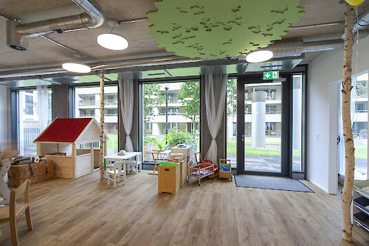 Kindergarten room with playhouse, large windows, round basic pendant luminaires and tree-shaped ceiling elements.