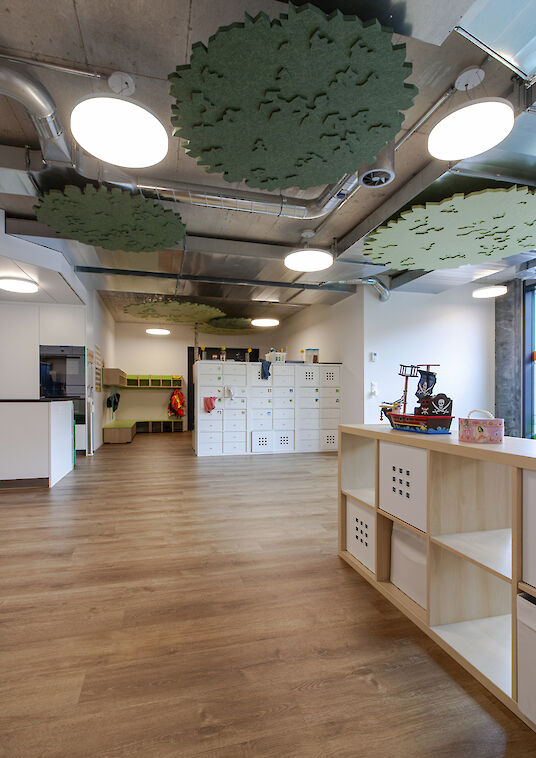 Open nursery room with wooden shelves, round basic pendant luminaires and green, tree-shaped ceiling elements.