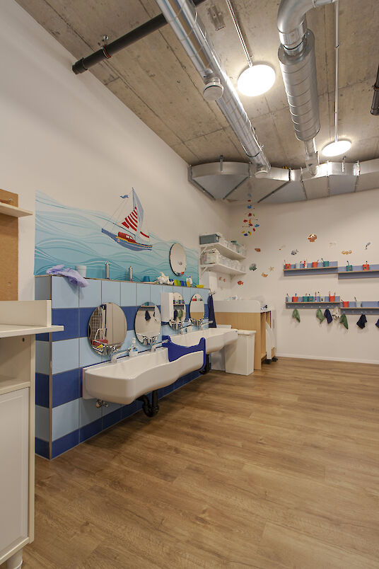 Child-friendly washroom with nautical wall design, several washbasins, wooden floor and round basic pendant luminaires.