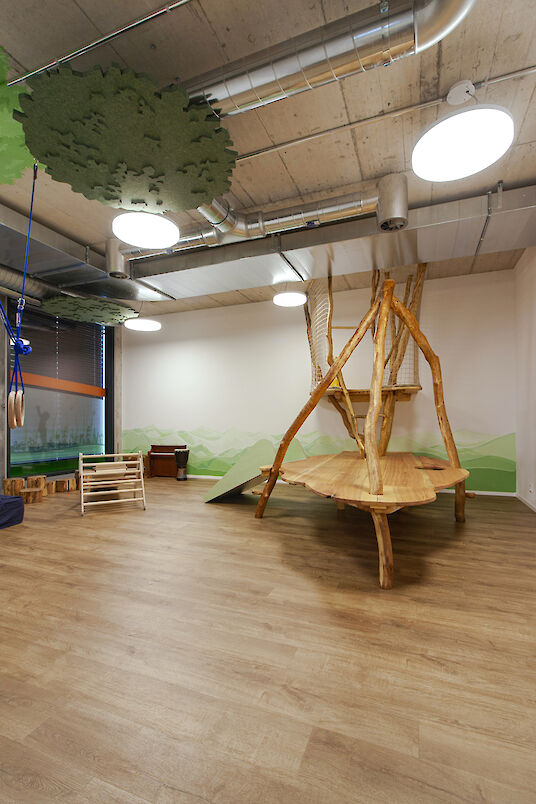 Playroom with wooden climbing landscape, round basic pendant luminaires and green tree-shaped ceiling elements.