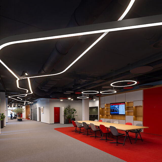 The interior of a modern savings bank: ceiling installations with curved Liquid Line light lines and circular pendant luminaires. The room has an open design, with red carpet highlights, a large conference table and decorative plants that create a harmonious atmosphere.