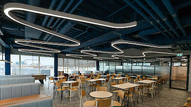 Canteen with curved Liquid Line LED light lines, open ceiling structures and wooden chairs at round tables.