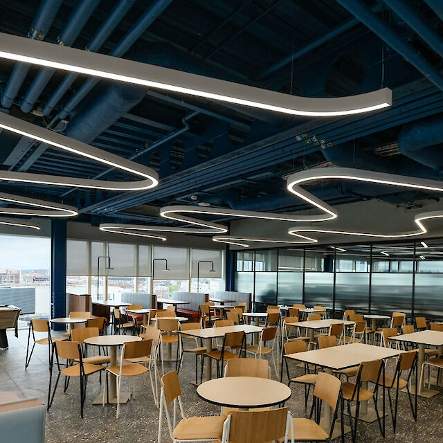 Canteen with curved Liquid Line LED light lines, open ceiling structures and wooden chairs at round tables.