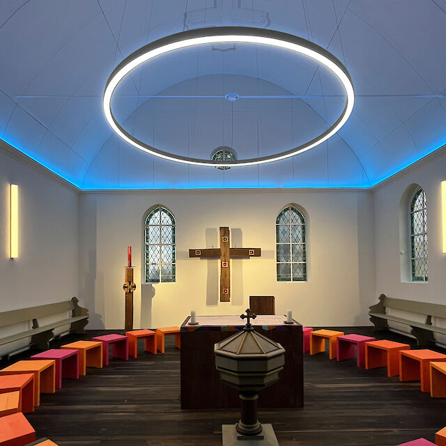 Modern illuminated chapel with a large, luminous ring and colourful stools in a semi-circle.