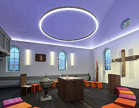 Capilla luminosa con un gran anillo de luces LED, taburetes de colores y una sencilla cruz de madera en la pared.