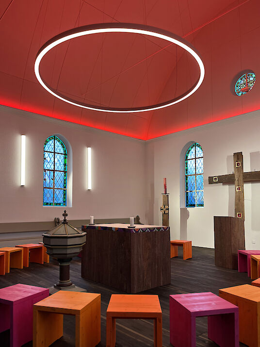 Capilla con lámpara de techo roja, gran anillo LED, taburetes de colores y sencilla cruz de madera
