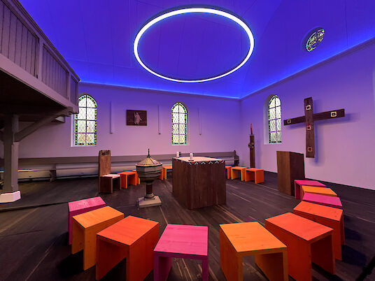 Capilla con techo iluminado en azul, gran anillo de LED y taburetes de colores en semicírculo