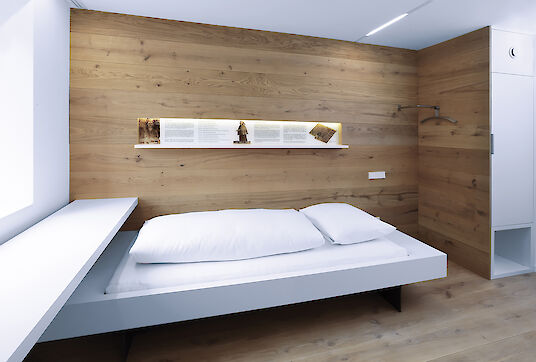 Schlicht eingerichtetes Hotelzimmer mit Holzverkleidung, weißem Bett, Ausstellungsnische und Matric Einbauleuchten.