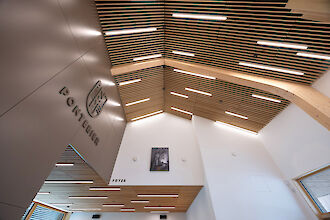 Interior view of a modern foyer with a high ceiling made of wooden slats, illuminated by narrow, linear LED lights. The wall displays the emblem and the word ‘Pontebier’, and below that the area is labelled ‘Foyer’. A work of art with a black and white tree motif hangs on the white wall, and large windows let daylight into the room.