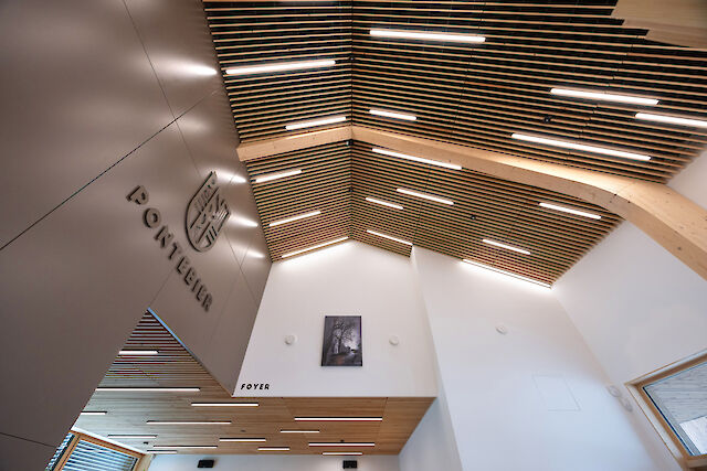 Interior view of a modern foyer with a high ceiling made of wooden slats, illuminated by narrow, linear LED lights. The wall displays the emblem and the word ‘Pontebier’, and below that the area is labelled ‘Foyer’. A work of art with a black and white tree motif hangs on the white wall, and large windows let daylight into the room.