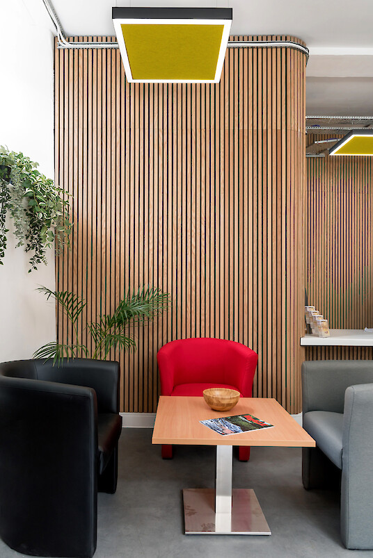 Coin salon avec fauteuils, table, plantes, paroi en lamelles de bois et suspension carrée avec panneau acoustique