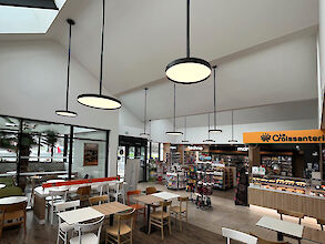 Interior view of a modern petrol station shop with café area. Several round, black pendant luminaires from Lightnet's Basic G3 series hang at different heights from the high white ceiling and illuminate the room evenly. Large window fronts and a glazed skylight allow daylight to flood in. In the foreground are tables and chairs in wood and white tones, while a sales area with shelves and a bakery counter with yellow signage can be seen in the background.