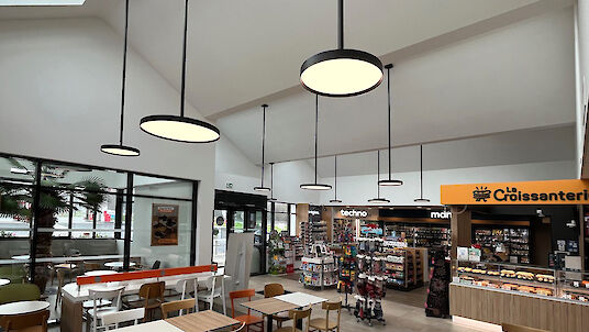 Interior view of a modern petrol station shop with café area. Several round, black pendant luminaires from Lightnet's Basic G3 series hang at different heights from the high white ceiling and illuminate the room evenly. Large window fronts and a glazed skylight allow daylight to flood in. In the foreground are tables and chairs in wood and white tones, while a sales area with shelves and a bakery counter with yellow signage can be seen in the background.