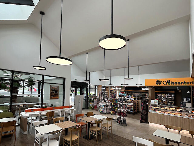 Innenansicht eines modernen Tankstellen-Shops mit Cafébereich. Mehrere runde, schwarze Pendelleuchten der Serie Basic G3 von Lightnet hängen in unterschiedlichen Höhen von der weißen, hohen Decke und beleuchten den Raum gleichmäßig. Große Fensterfronten und ein verglastes Oberlicht lassen Tageslicht einfallen. Im Vordergrund stehen Tische und Stühle in Holz- und Weißtönen, während im Hintergrund ein Verkaufsbereich mit Regalen und einer Bäckerei-Theke mit gelber Beschilderung zu sehen ist.