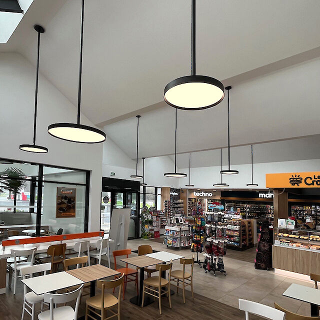 Innenansicht eines modernen Tankstellen-Shops mit Cafébereich. Mehrere runde, schwarze Pendelleuchten der Serie Basic G3 von Lightnet hängen in unterschiedlichen Höhen von der weißen, hohen Decke und beleuchten den Raum gleichmäßig. Große Fensterfronten und ein verglastes Oberlicht lassen Tageslicht einfallen. Im Vordergrund stehen Tische und Stühle in Holz- und Weißtönen, während im Hintergrund ein Verkaufsbereich mit Regalen und einer Bäckerei-Theke mit gelber Beschilderung zu sehen ist.