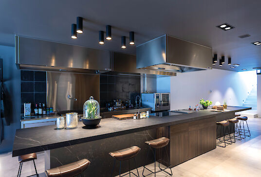 Moderna cocina de exposición con isla de cocción oscura, taburetes de bar, paneles de acero inoxidable y focos LED Midpoint.
