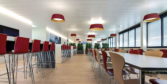 Cantina con mesas altas y bajas, sillas de colores y luminarias suspendidas rojas Beam Me Up.