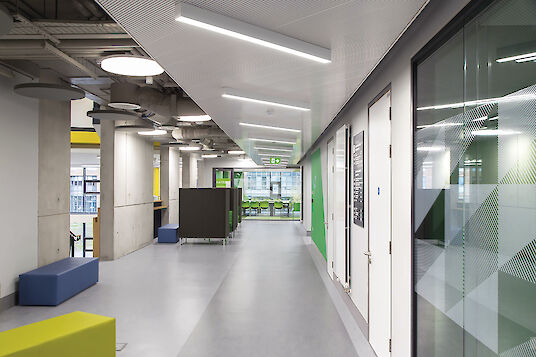 Hallway with colourful seating, exposed concrete, glass walls, linear Matric surface-mounted luminaires and Basic pendant luminaires.