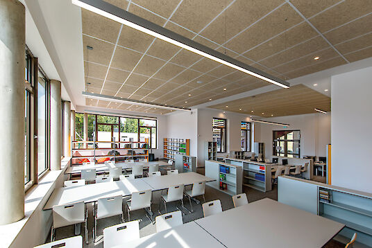 Light-flooded learning room with white tables, shelves, acoustic ceiling and linear Matric pendant luminaires.