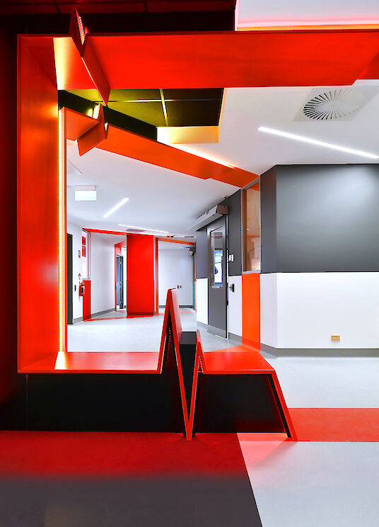 Corridor with red wall and ceiling surfaces, zigzag seating object and linear Matric recessed luminaires.