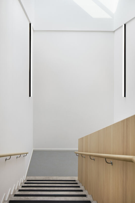 Staircase with wooden handrail and two vertically arranged Matric wall luminaires on white walls.