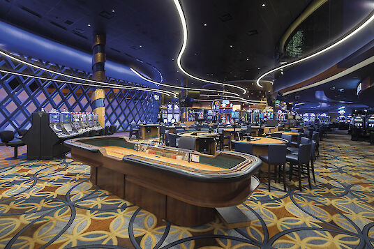 Casino with gaming tables, slot machines, floral patterned carpet and curved Liquid Line ceiling luminaire.