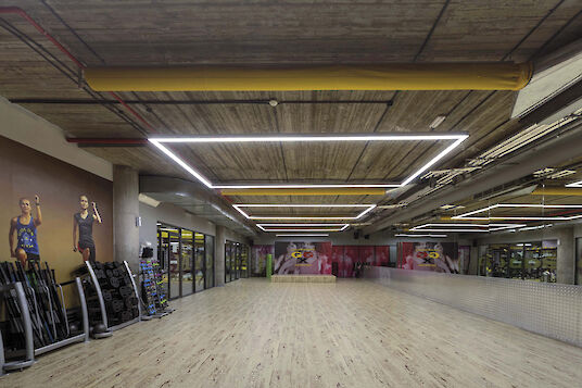 Amplia sala de fitness con suelo de madera, pared de espejos y luminarias Matric cuadradas en el techo de hormigón.