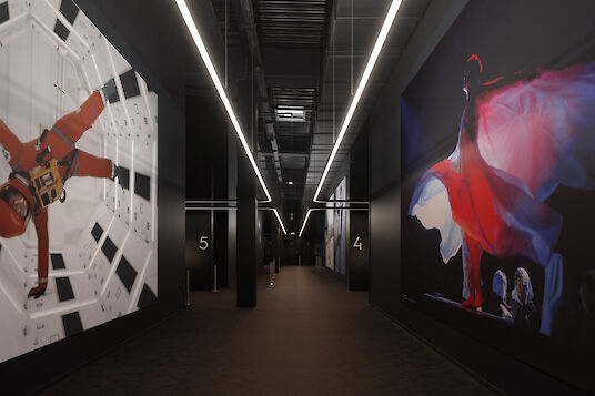 Cinema hallway with large-format film graphics, dark walls and linear Liquid Line pendant luminaires on the ceiling.