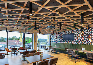 Modern restaurant with geometric wooden ceiling, midpoint lights, green upholstered bench and large window front to the terrace.