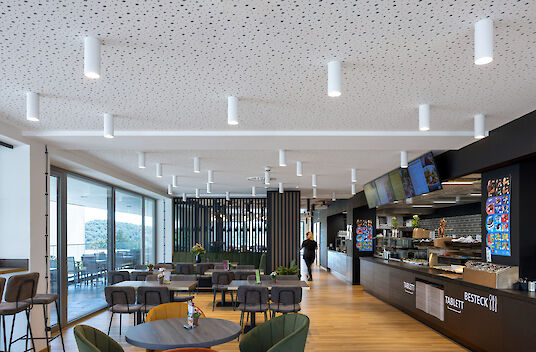 Cafeteria with wooden floor, self-service counter, round tables and white ‘Midpoint’ surface-mounted spotlights.