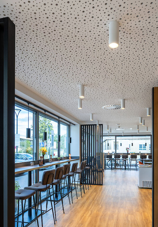 Cafeteria with window front, wooden floor, high tables and white ‘Midpoint’ surface-mounted spotlights on the ceiling.