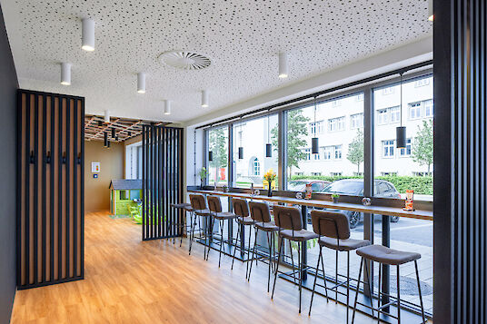  Cafeteria area with window front, bar tables, wooden floor and white ‘Midpoint’ surface-mounted spotlights on the ceiling.