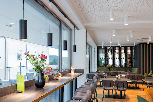 Cafeteria with window front, bar table, ‘Midpoint’ surface-mounted spotlights on the ceiling and brown chairs.