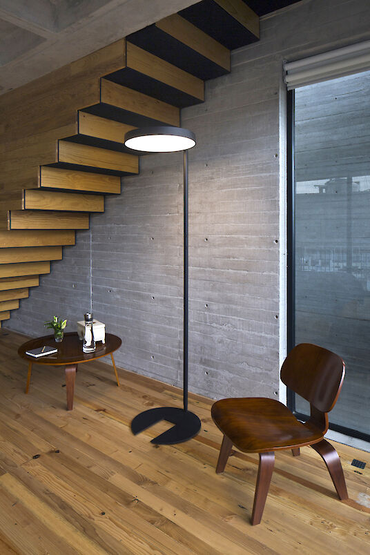 Modern floor lamp in front of an exposed concrete wall, next to a wooden chair, side table and open wooden staircase.