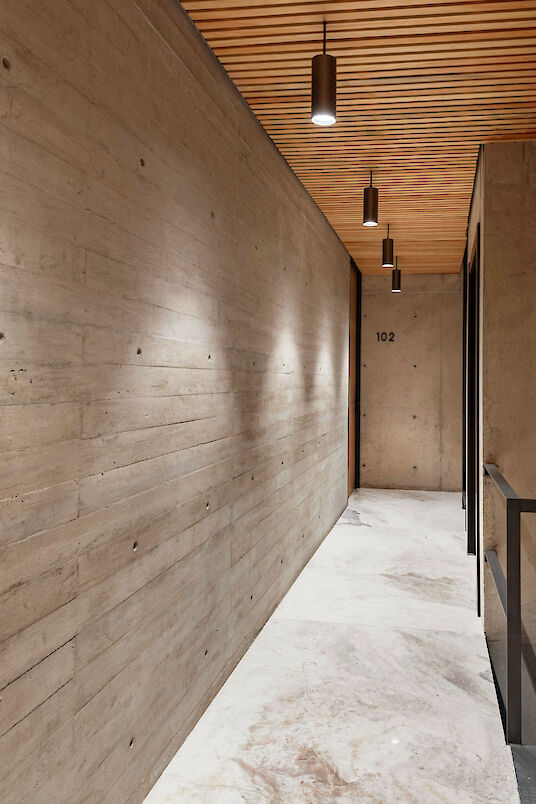Pasillo estrecho con paredes de hormigón visto, techo de listones de madera, focos cilíndricos en el techo y suelo de mármol claro.
