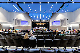 Auditorium with people, curved ceiling, integrated light lines and illuminated stage.
