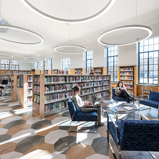 Biblioteca luminosa con luminarias suspendidas Ringo Star, estanterías, zona de lectura y suelo geométrico.