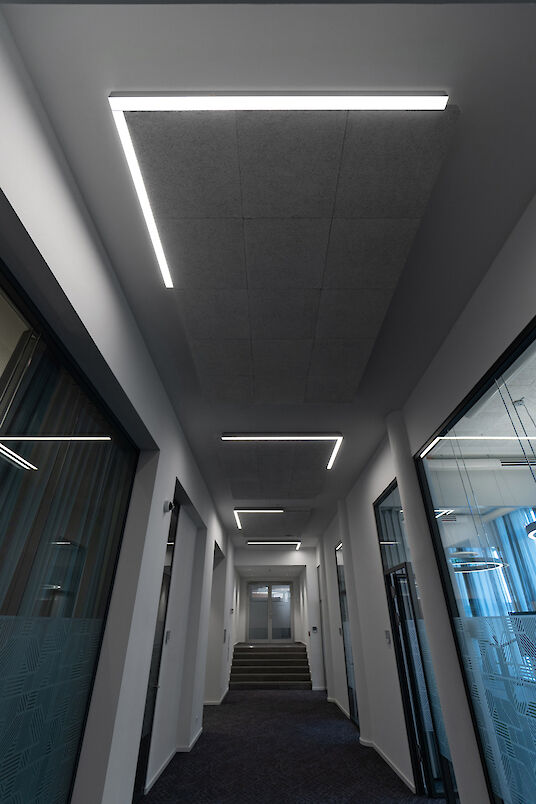 Vista de un moderno pasillo de oficinas con paredes blancas, despachos acristalados a ambos lados y un techo con paneles acústicos. Las luminarias lineales Matric A3 estructuran el techo a intervalos regulares y crean una iluminación uniforme y sin deslumbramientos del pasillo.