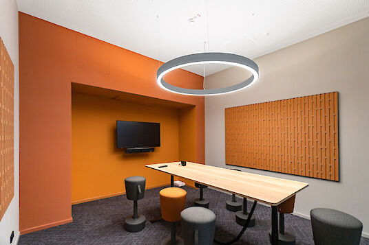 Pequeña sala de reuniones con paredes de color naranja, paneles de pared acústicamente eficaces y una mesa rectangular de madera con taburetes regulables en altura. Una luminaria circular Ringo Star P3 está suspendida en el centro por encima de la mesa y proporciona una luz uniforme y sin deslumbramientos. En la pared trasera hay una pantalla para presentaciones.
