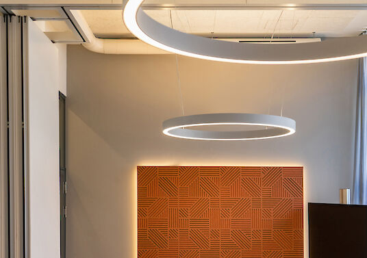 Vista detallada de una moderna sala de reuniones con dos luminarias Ringo Star P3 suspendidas en forma de anillo. Al fondo, un panel acústico de gran formato en naranja cálido con estructura gráfica, suavemente retroiluminado. El techo presenta instalaciones visibles y un lenguaje arquitectónico claro y minimalista.