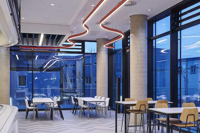 Interior de un moderno restaurante de empresa con luminarias Liquid Line curvadas y ventanas que van del suelo al techo.