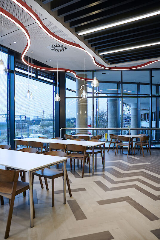 Zona de comedor con mesas blancas, sillas de madera e iluminación Liquid Line curvada frente a grandes ventanales.