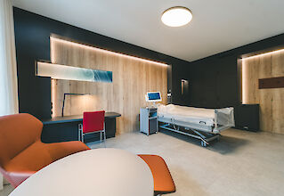 Modern eingerichtetes Patientenzimmer mit Pflegebett, Holzvertäfelung, Schreibtisch und blendfreier Deckenleuchte