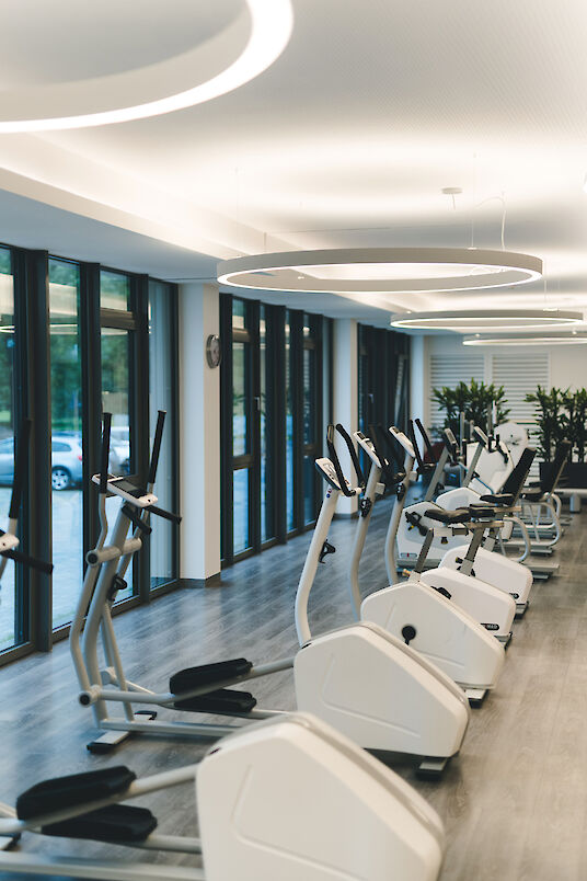 Reihe weißer Ergometer vor bodentiefen Fenstern, darüber ringförmige Pendelleuchten in heller Trainingszone